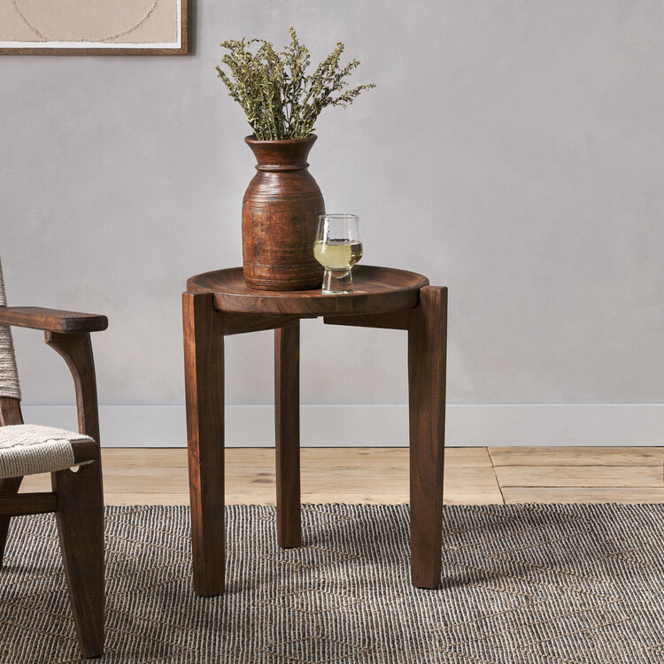 Dark brown acacia wood side table with a circular top and three legs holding a glass a vase